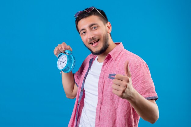 Jeune beau mec voyageur tenant un réveil à la recherche d'un sourire positif et heureux montrant les pouces vers le haut debout sur fond bleu