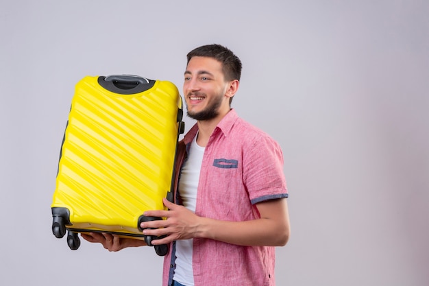 Jeune beau mec voyageur tenant valise à côté positif et heureux souriant debout sur fond blanc