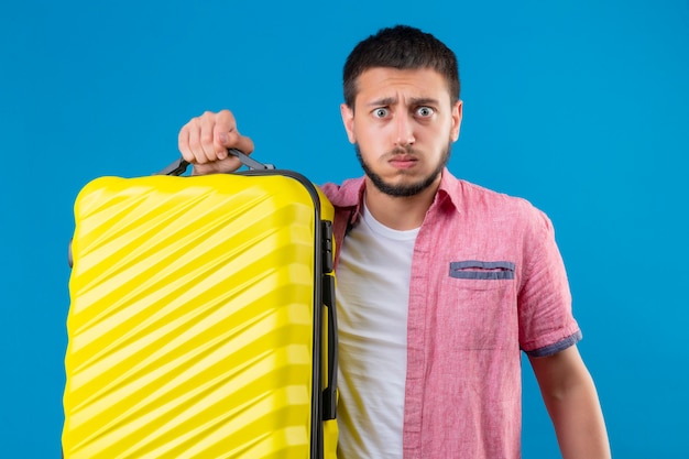 Jeune beau mec voyageur tenant valise à l'ennui et gêné de souffler les joues debout sur fond bleu