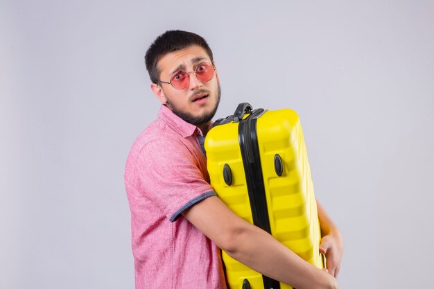 Jeune beau mec voyageur tenant valise regardant la caméra confondu avec une expression triste sur le visage debout sur fond blanc