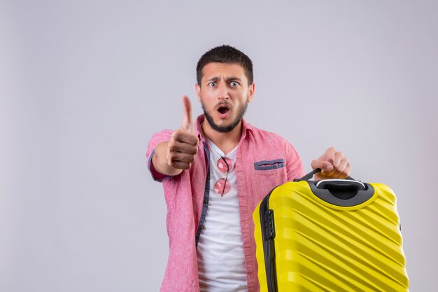 Jeune beau mec voyageur tenant valise regardant la caméra surpris et étonné montrant les pouces vers le haut debout sur fond blanc