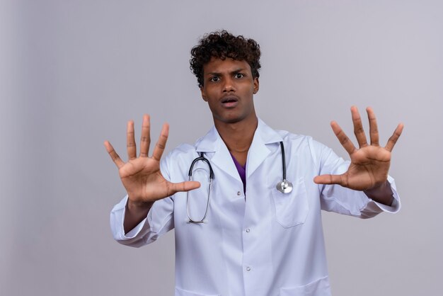 Un jeune beau médecin à la peau foncée avec des cheveux bouclés portant une blouse blanche avec un stéthoscope se tenant la main en arrêt ou assez de geste sur un fond blanc