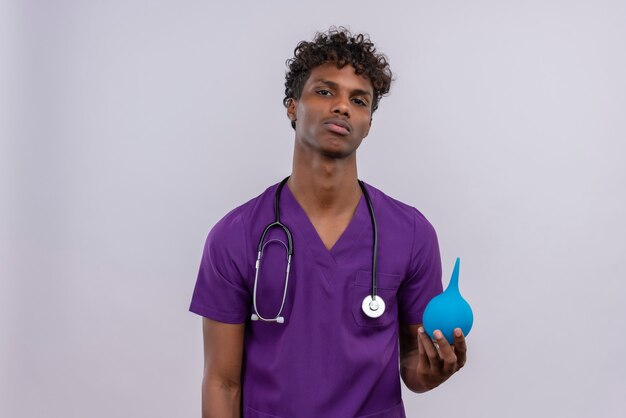 Un jeune beau médecin à la peau foncée avec des cheveux bouclés portant un uniforme violet avec stéthoscope tout en tenant un lavement