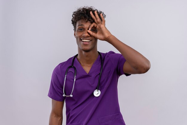 Un jeune beau médecin à la peau sombre aux cheveux bouclés portant un uniforme violet avec stéthoscope furtivement à travers un trou formé avec son pouce et son index