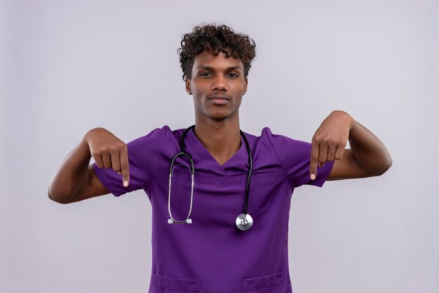 Un jeune beau médecin à la peau sombre aux cheveux bouclés portant l'uniforme violet avec stéthoscope pointant vers le haut avec l'index