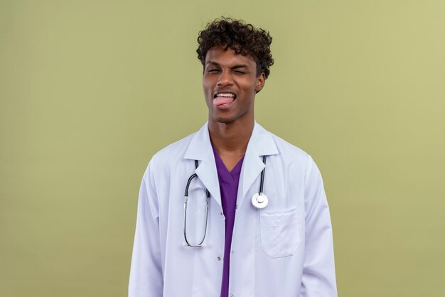 Un jeune beau médecin à la peau sombre avec des cheveux bouclés portant un blouse blanche avec un stéthoscope montrant sa langue sur un espace vert