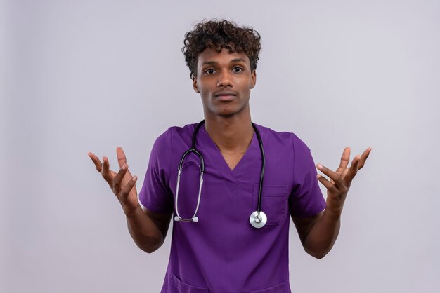 Un jeune beau médecin à la peau sombre avec des cheveux bouclés portant l'uniforme violet avec stéthoscope ouvrant les mains avec une expression confuse