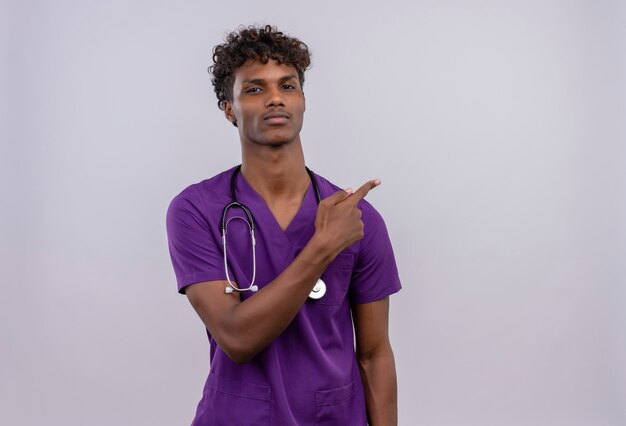 Un jeune beau médecin à la peau sombre avec des cheveux bouclés portant un uniforme violet avec stéthoscope tout en pointant avec l'index vers le haut