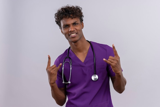 Un jeune beau médecin à la peau sombre confiant avec des cheveux bouclés portant l'uniforme violet avec stéthoscope se tenant la main dans le geste de la roche