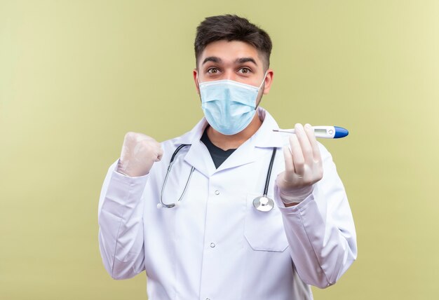 Jeune beau médecin portant un masque médical bleu robe médicale blanche gants médicaux blancs et stéthoscope tenant joyeusement thermomètre électronique heureux pour les résultats de la température