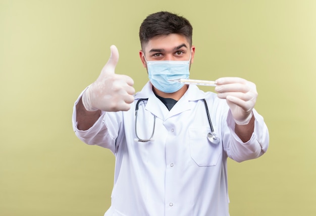 Jeune beau médecin portant un masque médical bleu robe médicale blanche gants médicaux blancs et stéthoscope tenant un thermomètre faisant des pouces heureux debout sur un mur kaki