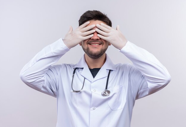 Jeune beau médecin portant une robe médicale blanche, des gants médicaux blancs et un stéthoscope jouant à cache-cache et malade heureusement debout sur un mur blanc