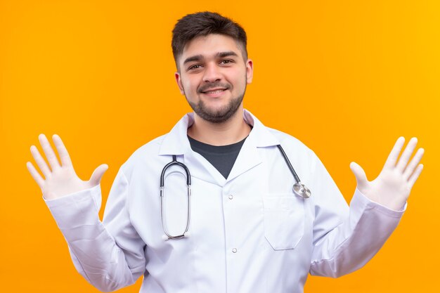 Jeune beau médecin portant une robe médicale blanche, des gants médicaux blancs et un stéthoscope souriant bras d'ouverture disant Salut debout sur un mur orange