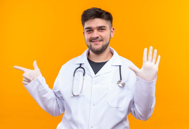 Jeune beau médecin portant une robe médicale blanche, des gants médicaux blancs et un stéthoscope souriant joyeusement montrant cinq signes avec les doigts et pointant sur la droite debout sur orange