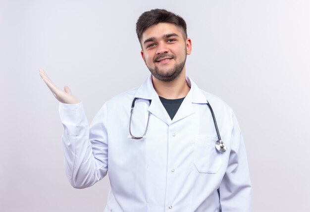 Jeune beau médecin portant une robe médicale blanche, des gants médicaux blancs et un stéthoscope souriant montrant quelque chose avec la main droite debout sur un mur blanc