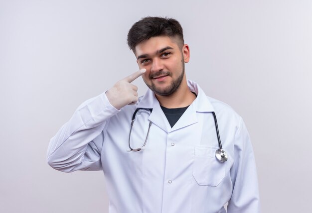 Jeune beau médecin portant une robe médicale blanche, des gants médicaux blancs et un stéthoscope souriant pointant sur son nez debout sur un mur blanc