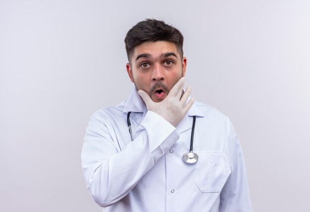 Jeune beau médecin portant une robe médicale blanche, des gants médicaux blancs et un stéthoscope à la surprise tenant sa mâchoire avec la main debout sur un mur blanc