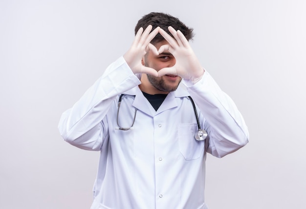 Jeune beau médecin portant une robe médicale blanche, des gants médicaux blancs et un stéthoscope à la timidement montrant signe d'amour avec les mains debout sur un mur blanc