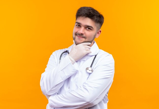 Jeune beau médecin portant une robe médicale blanche, des gants médicaux blancs et un stéthoscope