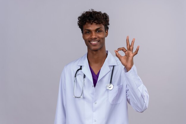 Un jeune beau médecin de sexe masculin à la peau foncée excité avec des cheveux bouclés portant une blouse blanche avec stéthoscope faisant signe ok avec les doigts