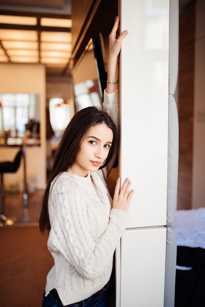 Jeune beau modèle tendre avec de grands yeux lèvres rouges et de longs cheveux bruns en pull blanc et un jean bleu restent dans la chambre chez elle