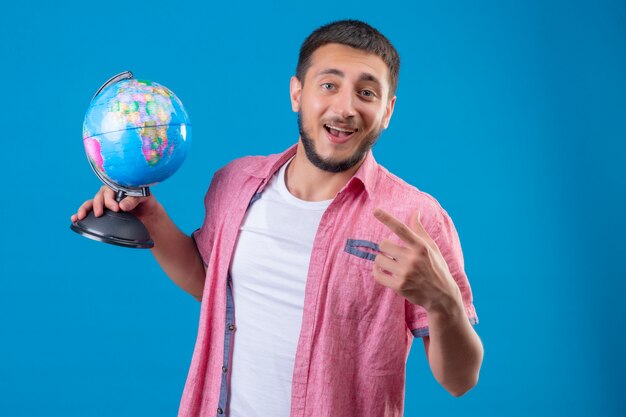 Jeune beau voyageur guy holding globe souriant avec visage heureux pointant avec le doigt sur le côté debout sur fond bleu