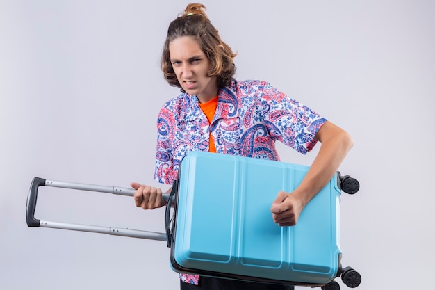 Jeune beau voyageur guy holding valise à l'aide d'une guitare à la recherche avec une expression de colère debout sur fond blanc