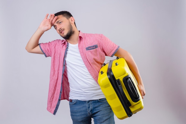 Jeune beau voyageur guy holding valise à côté ennuyé et fatigué avec une expression triste sur le visage debout sur fond blanc