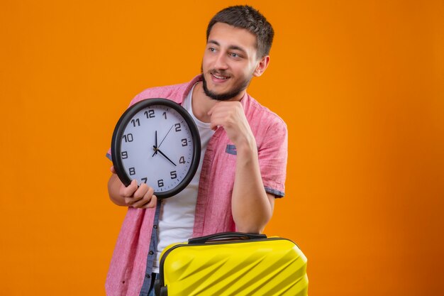 Jeune beau voyageur guy tenant horloge à côté avec la main sur le menton pensant positif valise d'esprit debout sur fond orange