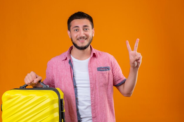 Jeune beau voyageur guy tenant valise regardant la caméra avec un visage heureux souriant montrant le numéro deux ou signe de la victoire debout sur fond orange