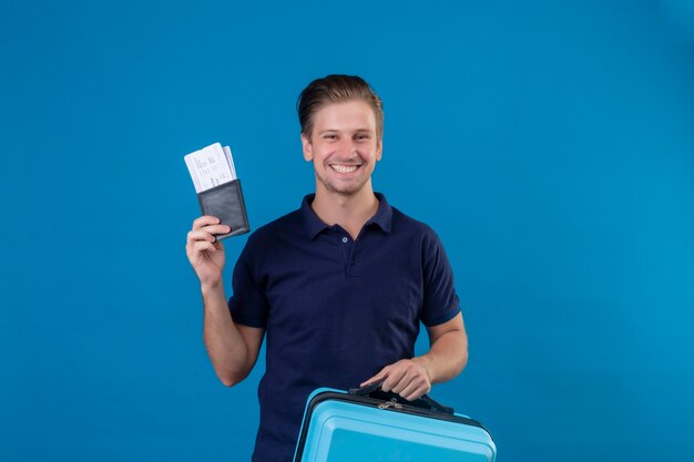 Jeune beau voyageur homme tenant des billets d'avion heureux et positif regardant la caméra avec un grand sourire sur le visage debout sur fond bleu