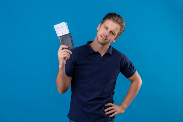 Jeune beau voyageur homme tenant des billets d'avion heureux et positif regardant la caméra avec un sourire confiant sur le visage debout sur fond bleu