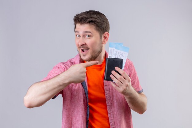 Jeune beau voyageur homme tenant des billets d'avion pointant avec le doigt pour eux souriant joyeusement positif et heureux debout sur un mur blanc