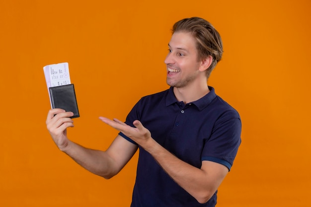 Jeune beau voyageur homme tenant des billets d'avion présentant le bras de la main à eux avec le sourire sur le visage sur fond orange