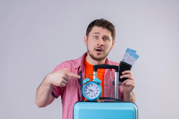 Jeune beau voyageur homme tenant valise bleue et billets d'avion avec réveil confus et déçu debout sur un mur blanc