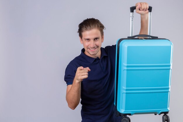 Jeune beau voyageur homme tenant valise à la caméra à doigt pointé confiant souriant debout sur fond blanc