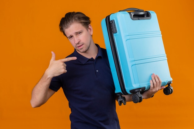 Jeune beau voyageur homme tenant valise à la recherche de doigt pointé confiant debout sur fond orange