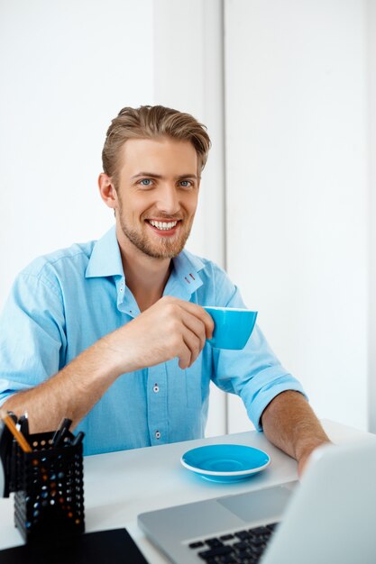 Jeune bel homme d'affaires confiant gai assis à table travaillant sur ordinateur portable, boire du café. souriant. Intérieur de bureau moderne blanc