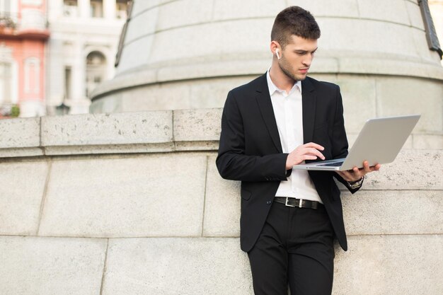 Jeune bel homme d'affaires en costume noir classique et chemise blanche avec des écouteurs sans fil debout pensivement travaillant sur un ordinateur portable tout en passant du temps à l'extérieur