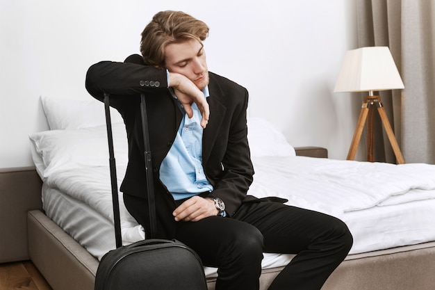 Jeune bel homme d'affaires en costume noir s'endort avec une valise dans la chambre d'hôtel après un long voyage en avion en mission commerciale.