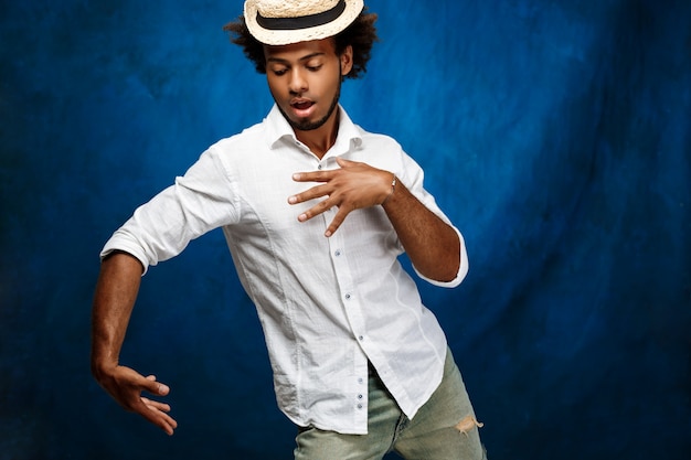 Jeune bel homme africain en chapeau dansant sur le mur bleu.