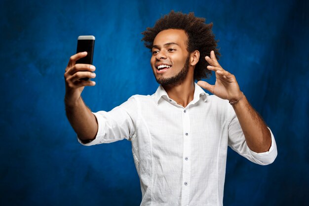 Jeune bel homme africain faisant selfie sur mur bleu.