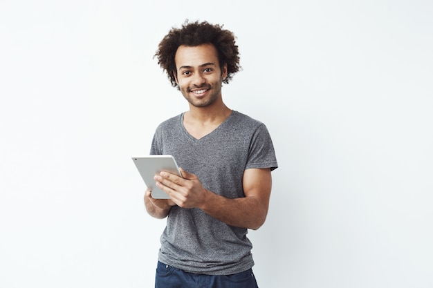 Jeune bel homme africain souriant tenant une tablette argentée et jouer à des jeux ou en utilisant une application de réservation contre le mur blanc.