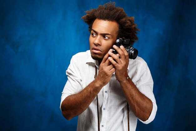 Jeune bel homme africain tenant un vieil appareil photo sur le mur bleu.