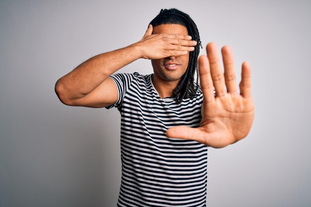 Jeune bel homme afro-américain avec des dreadlocks portant un t-shirt rayé et des lunettes couvrant les yeux avec les mains et faisant un geste d'arrêt avec une expression de tristesse et de peur Concept embarrassé et négatif