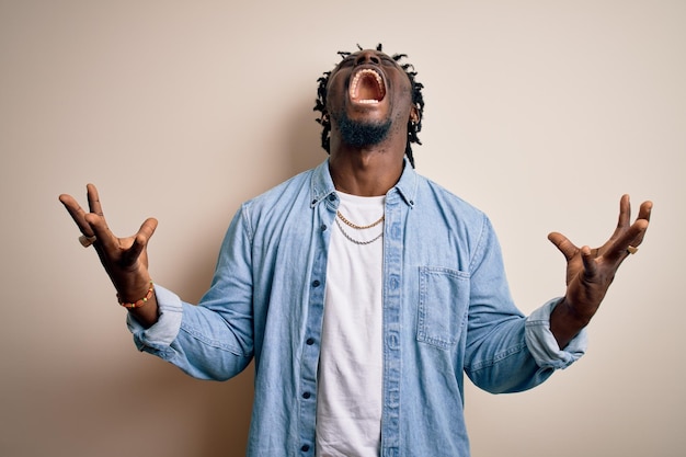 Jeune bel homme afro-américain portant une chemise en jean décontractée sur fond blanc fou et fou criant et hurlant avec une expression agressive et les bras levés Concept de frustration