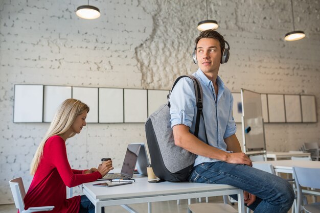 Jeune bel homme assis sur la table dans les écouteurs avec sac à dos dans le bureau de travail, boire du café,