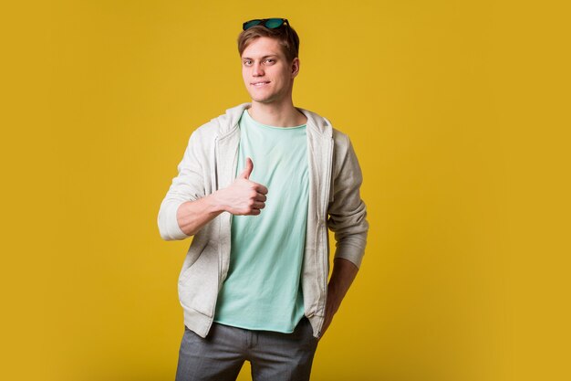 Jeune bel homme avec barbe debout sur un mur jaune souriant avec un visage heureux à la recherche et pointant vers le côté avec le pouce vers le haut.