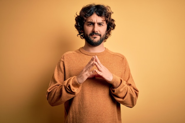 Jeune bel homme avec barbe portant un pull décontracté debout sur fond jaune Mains jointes et doigts croisés souriant détendu et joyeux Succès et optimiste