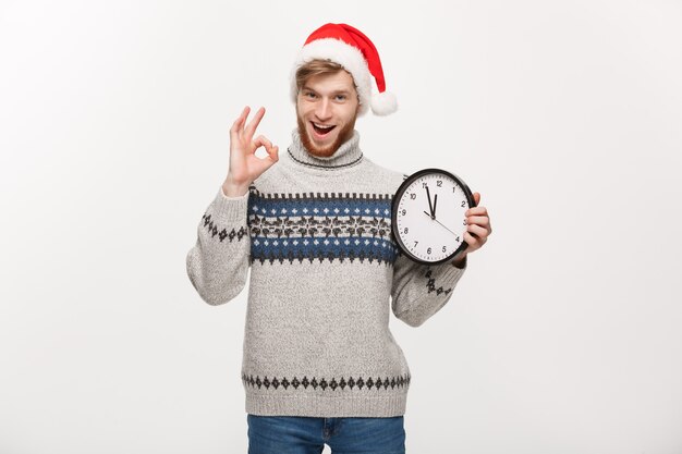 Jeune bel homme barbe en pull avec horloge blanche donnant signe ok.